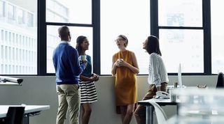 Four colleagues stood by a window holding coffee mugs and laughing